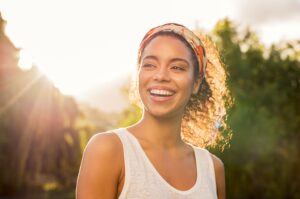 Woman in white tank top outside smiling with sunbeams behind her